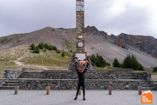 Smiling cyclist lifts his bike overhead at the summit of Col d'Izoard